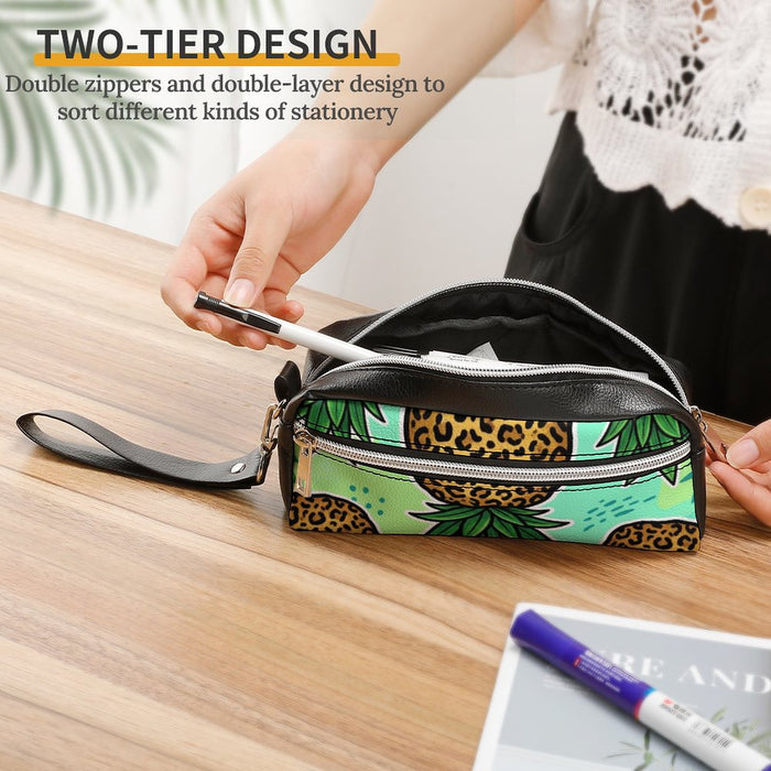 A two-tier pencil case with a green tropical leaf print and leopard print accents, shown on a wooden table with a person's hands inserting a pen into the top compartment. Text overlay at the top reads 'TWO-TIER DESIGN'.